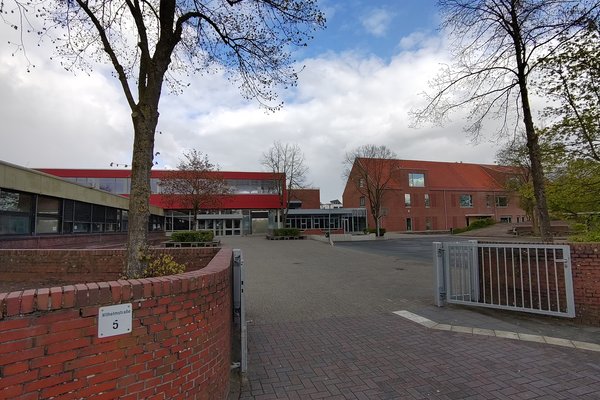 Im Vordergrund ein Tor in  einer Backsteinmauer. An der linken Seite der Mauer ein Schild "Wilhelmstraße 5". Im Hintergrund links ein heruntergekommener eingeschossiger Flachbau. Dahinter ein zweigeschossiges, moderner aussehendes Gebäude mit flachem Dach. Rechts hinten ein zweigeschossiger Neubau mit Satteldach.