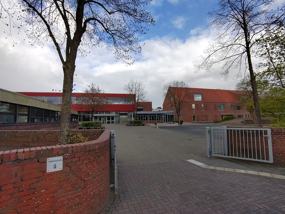 KGS Rastede Im Vordergrund ein Tor in einer Backsteinmauer. An der linken Seite der Mauer ein Schild "Wilhelmstraße 5". Im Hintergrund links ein heruntergekommener eingeschossiger Flachbau. Dahinter ein zweigeschossiges, moderner aussehendes Gebäude mit flachem Dach. Rechts hinten ein zweigeschossiger Neubau mit Satteldach.